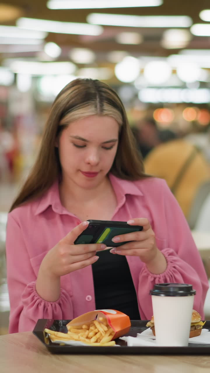 dama blanca en vestido rosa sentada en una mesa de madera en un restaurante ocupado, tomando una foto de su comida que consiste en una hamburguesa, papas fritas y una taza de café en una bandeja negra delante de ella