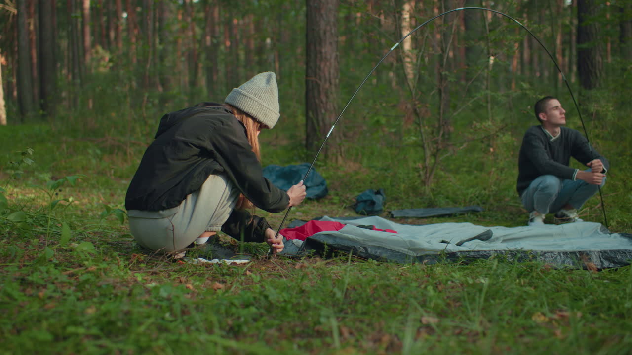 Man holds flexible tent pole while woman squats on forest floor securing her end and attaching tent fabric, surrounded by trees and gear, they cooperate peacefully during outdoor camping setup