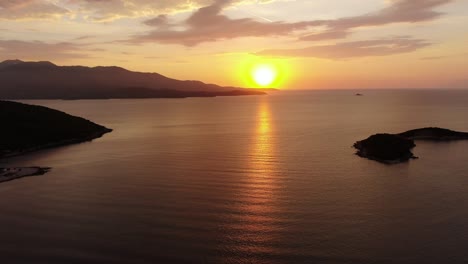 Drohnenansicht-In-Albanien,-Die-Bei-Sonnenuntergang-über-Blauem-Kristallklarem-Wasser-Fliegt,-Wobei-Die-Sonne-Am-Horizont-Mit-Kleinen-Inseln-In-Ksamil-Untergeht