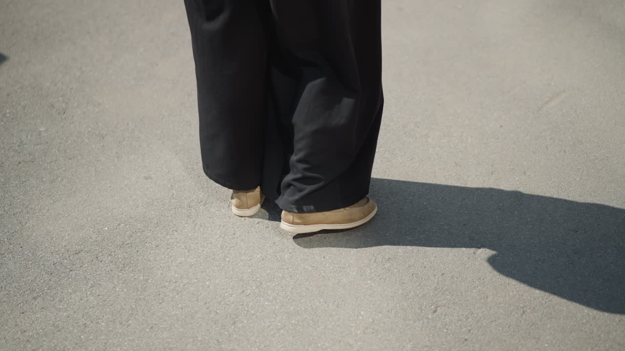Lower view of individual standing quietly on bright sunlit pavement wearing black wide pants and brown shoes, long shadow stretches across tarred ground
