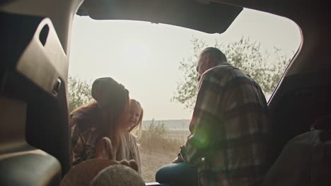 A-happy-man-with-gray-hair-in-a-plaid-shirt-sits-in-the-open-trunk-of-a-car-and-communicates-with-his-little-daughter-and-his-wife,-a-brunette-girl-in-a-plaid-shirt,-during-his-vacation-outside-the-city-in-the-summer