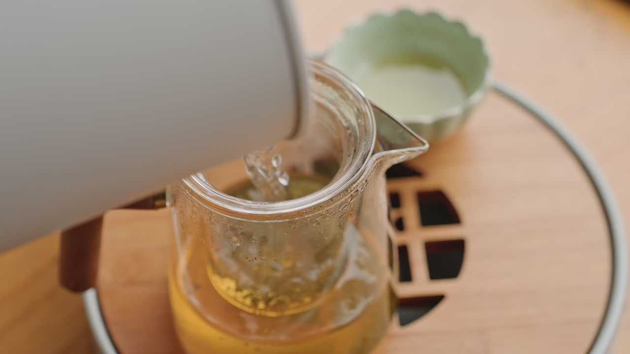 Vassoio dell'hotel con teiera in vetro in infusione, primo piano di un bollitore che versa acqua calda su foglie di tè verde, vapore delicato che sale, vassoio di servizio in legno, piccola tazza in ceramica nelle vicinanze, atmosfera di calda ospitalità, versamento lento.