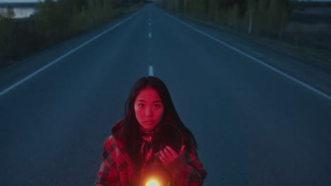 portrait of woman holding flickering lantern on empty road in evening