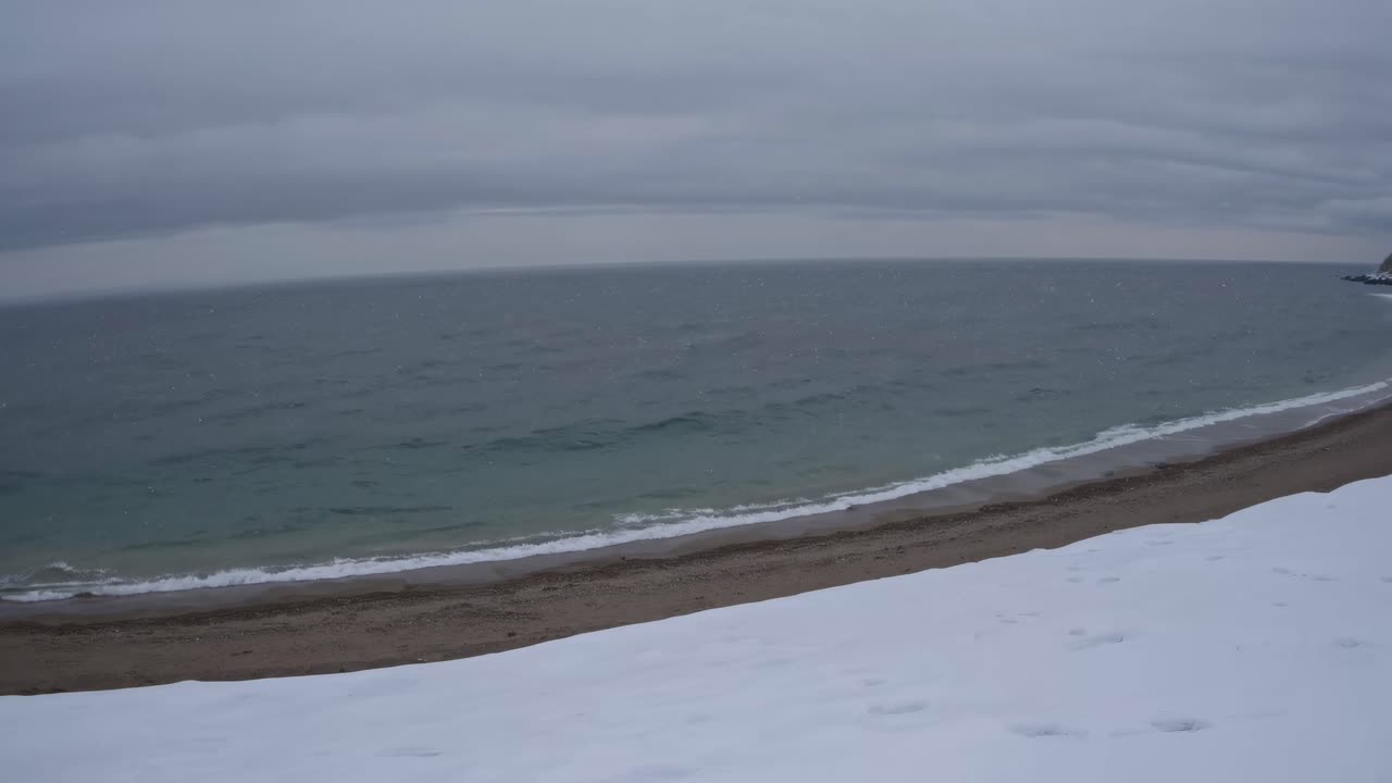 A wide-angle video captures a serene, snowy beach with gentle waves under a cloudy sky