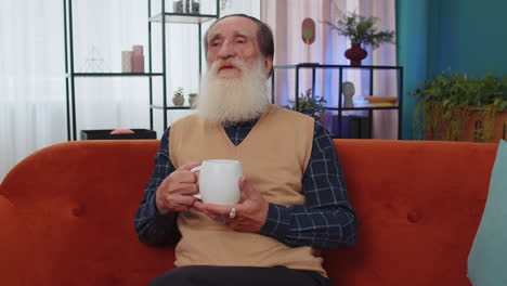 Attractive-smiling-senior-old-grandfather-man-drinking-a-cup-of-coffee-or-herbal-tea-at-home-couch