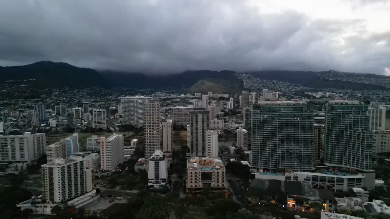 Veduta aerea dello skyline di Honolulu alle Hawaii, con grattacieli e hotel sotto un cielo drammatico e nuvoloso e montagne visibili sullo sfondo mentre la sera si avvicina alla città tropicale.