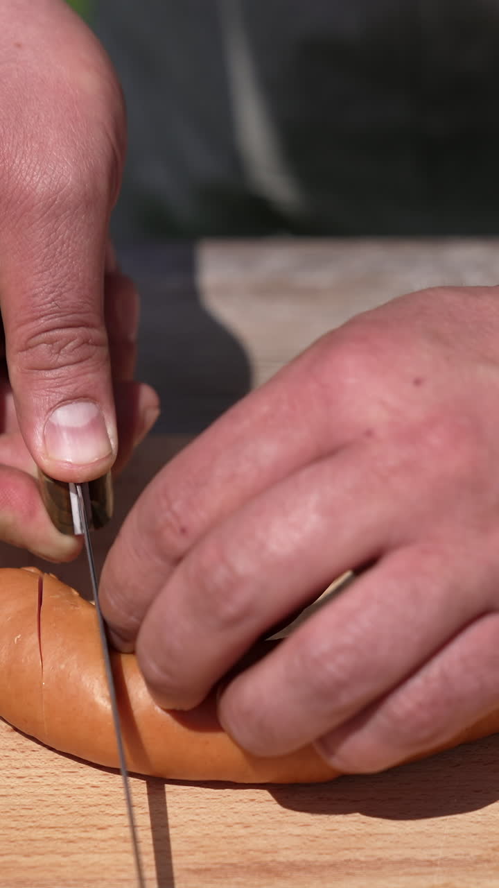 Cooking sausages in the open air. Man making an incision on sausages with a sharp knife before frying on a grill. Close-up. Vertical video