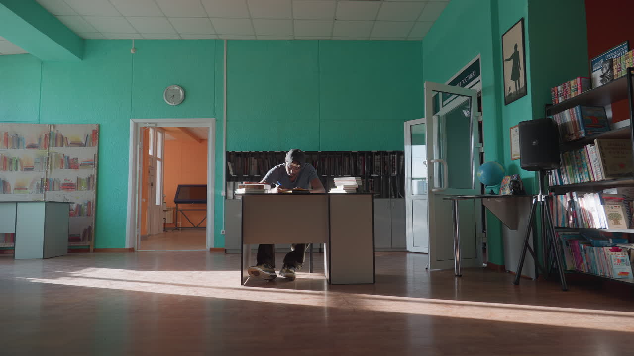 Wide view of young man wearing cap sitting at desk reading books in spacious brightly lit library with turquoise walls, wooden floor, open door, bookshelves, and scattered sunlight