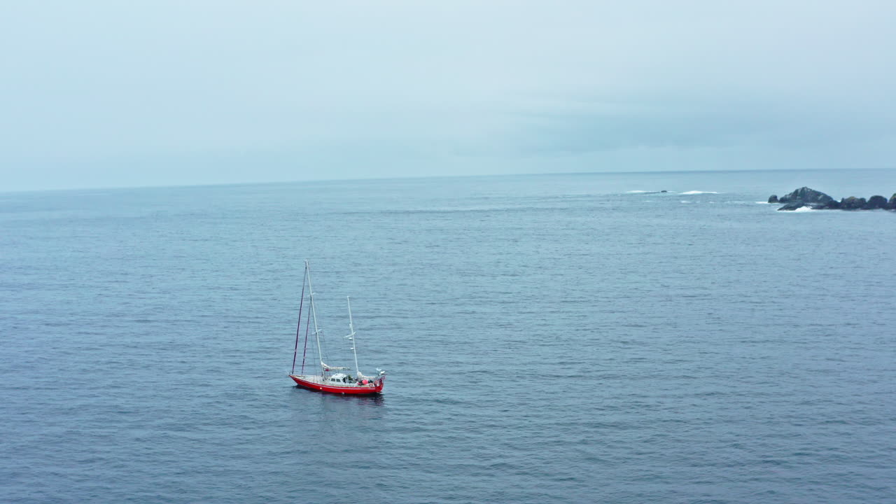 Solo sailboat drifts across open Beagle Channel in calm weather under a light blue sky, aerial orbit