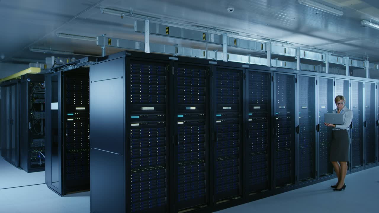 In Data Center: Female IT Technician Running Maintenance Programme on a Laptop, Controls Operational Server Rack Optimal Functioning. Modern High-Tech Telecommunications Operational Super Computer Database.