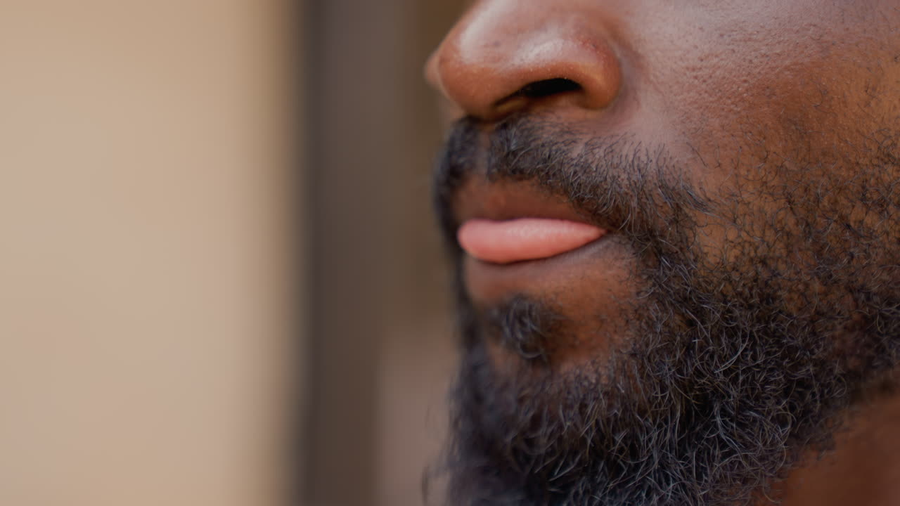 Primo piano di profilo di un uomo di colore con barba, che esprime tranquilla contemplazione e una barba incolta ben definita, dettaglio intimo della mascella e sottile movimento della bocca, sfondo neutro caldo, luce naturale, un'atmosfera spontanea e riflessiva.