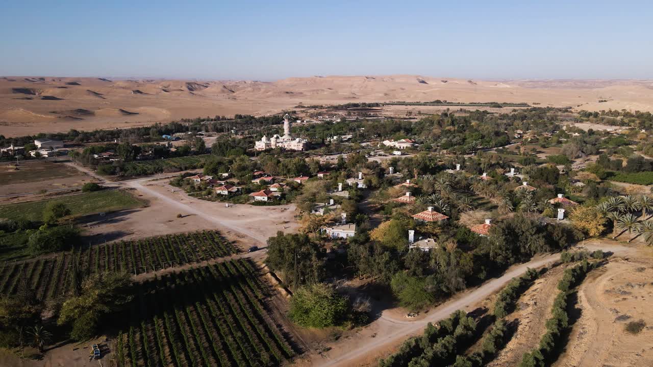 Wide view of Neot Smadar kibbutz with its iconic Art Center surrounded by agricultural fields, orchards, and desert hills under clear sky, drone slow orbiting