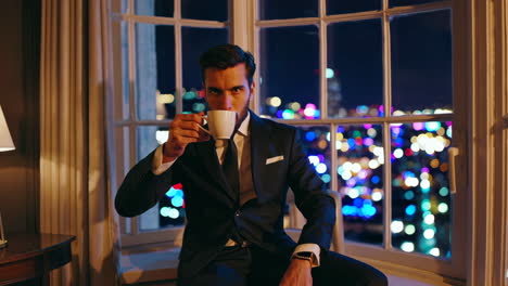 man in a suit drinking by a window with city night view