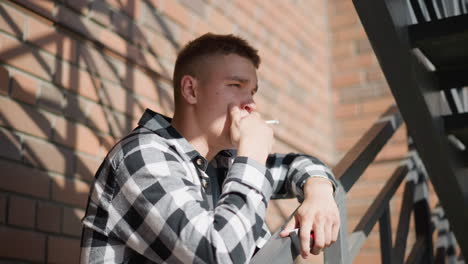 side view of student resting on iron railing smoking cigarette while gazing thoughtfully into distant urban scene with brick wall shadows metal staircase railing under warm sunlight