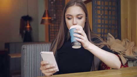 young pretty woman in casual clothing holding cup of coffee and using smartphone