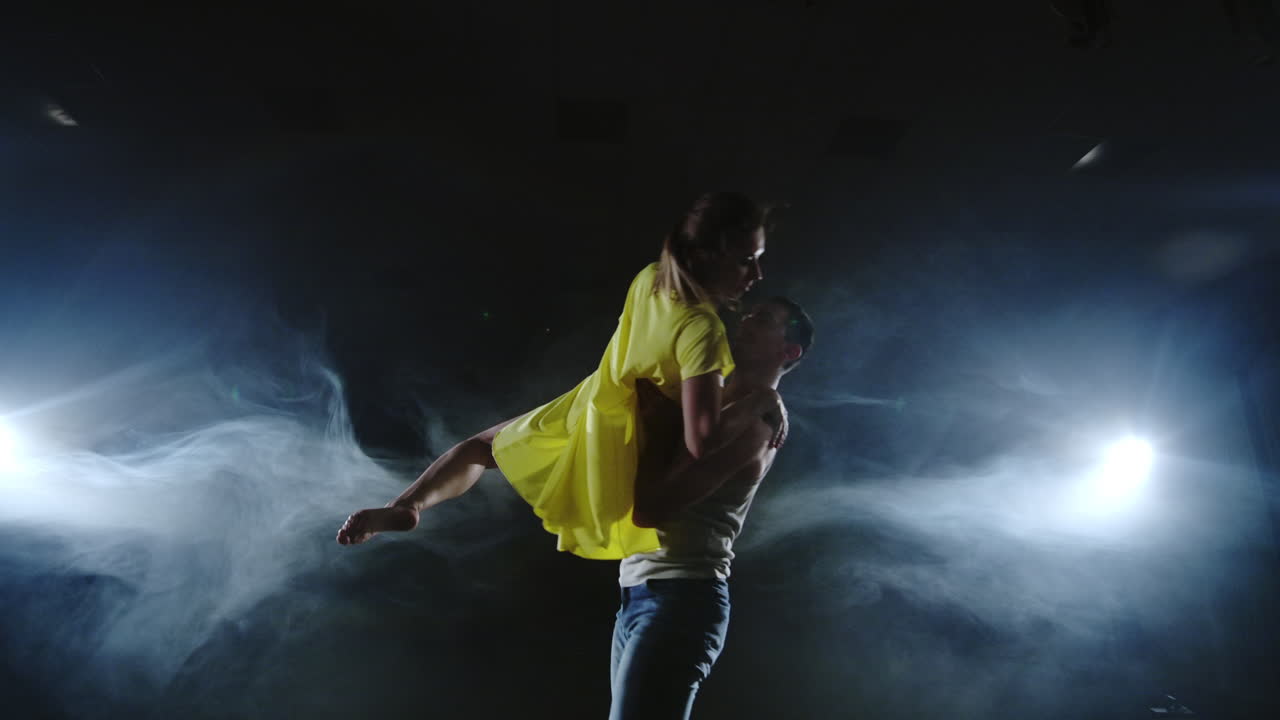a young girl in yellow dress and a man are running towards each other and doing acrobatic tricks in slow motion on stage in smoke and spotlights. camera is zooming in and out
