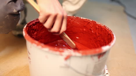 close up of a painter stirring with a wooden stick in a paint bucket full of red paint