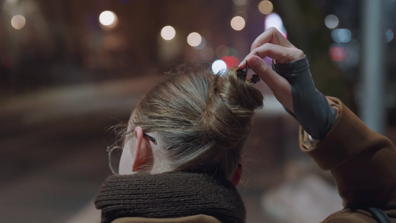Strada cittadina notturna, pendolare donna (vista di spalle) si ferma sotto luci bokeh per sciogliere lo chignon e raccogliere i capelli in una coda di cavallo, che indossa sciarpa e cappotto di lana, guanti senza dita visibili, neve sul marciapiede.