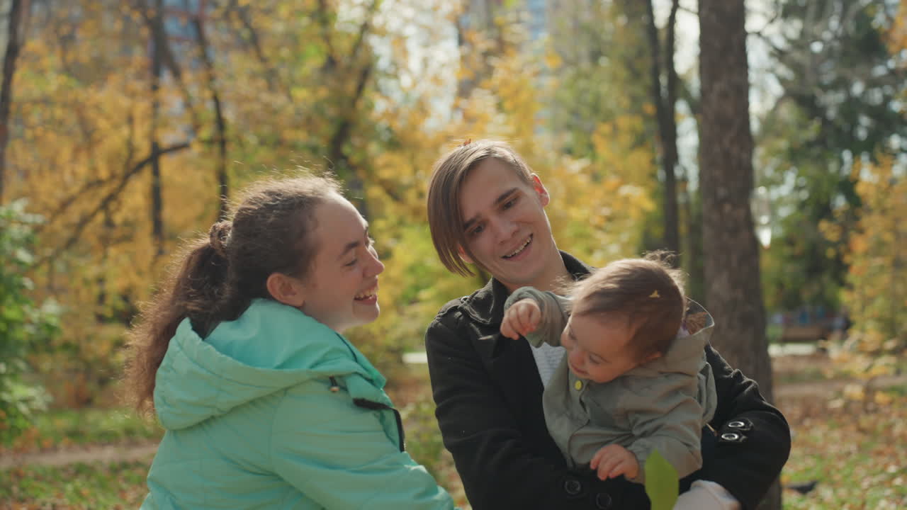 Herbstpark, Familie, weiße Mutter und Schwester spielen mit Baby, zärtliche Bindung im Freien zwischen Bäumen und goldenen Blättern, legere Mäntel und warmes Lächeln, Geschwisterinteraktion und sanfte Streicheleinheiten, friedlicher Tag