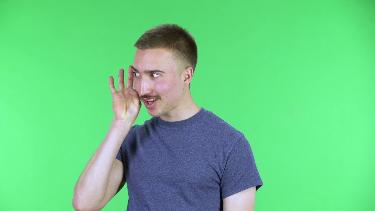 Ritratto di giovane uomo che tiene una mano vicino all'orecchio cercando di ascoltare notizie interessanti, esprimendo un concetto di comunicazione e pettegolezzo. Un uomo carino con i baffi in posa su schermo verde in studio. Primo piano.