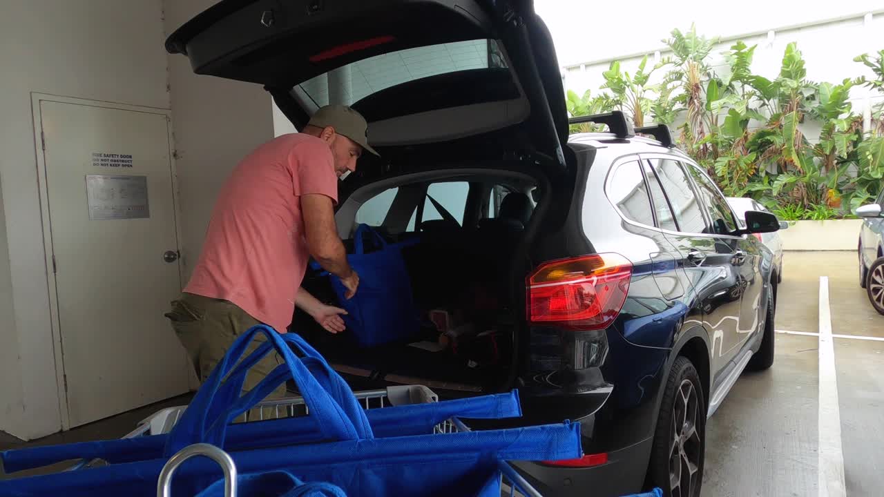 한 성숙한 남자가 쇼핑 카트를 풀고 식료품을 차 뒤에 싣고 있다
