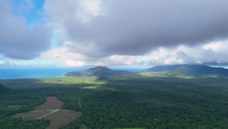 drone footage glides above dense rainforest and agricultural fields, revealing dramatic cloud shadows and sunlight over a tropical landscape with distant mountains