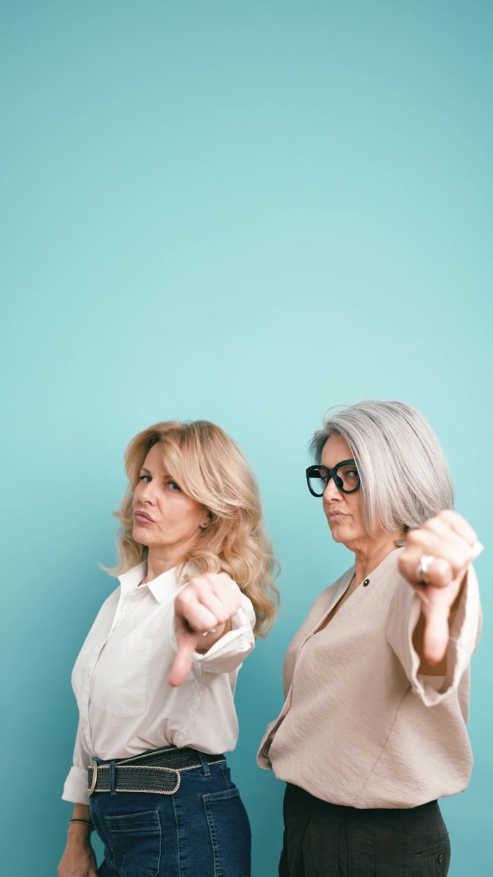 Two Mature Women Giving Thumbs Down Gesture Against Teal Background