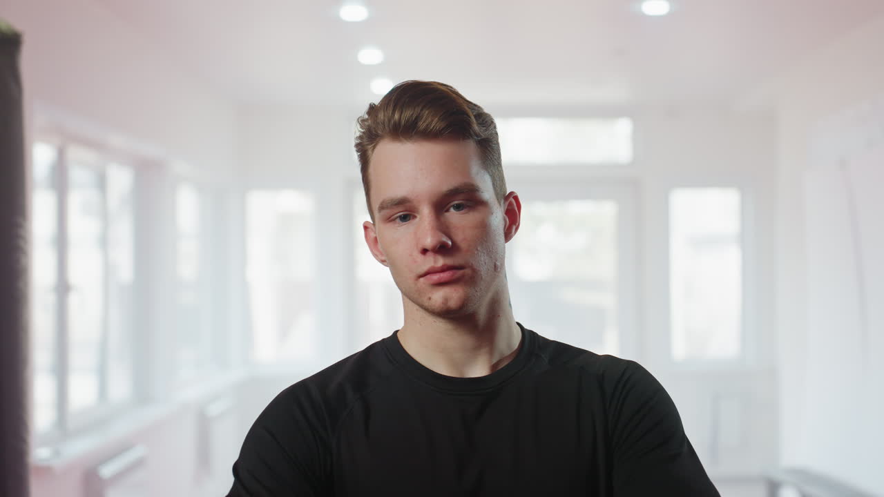 Athlete in black sportswear stands inside gym between punching bags looking down with focused serious posture, representing strength, preparation, discipline, and determination before combat session