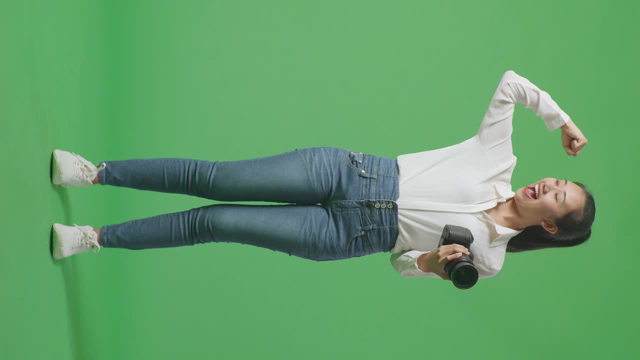 스튜디오에서 녹색 화면 배경에 서 있는 동안 사진을 찍고 그녀의 양팔을 구부리는 카메라를 사용하는 아시아 사진작가의 전체 몸