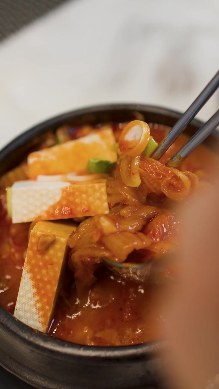 Sendok logam menyendok kimchi dan tahu dari mangkuk batu panas yang mendidih.