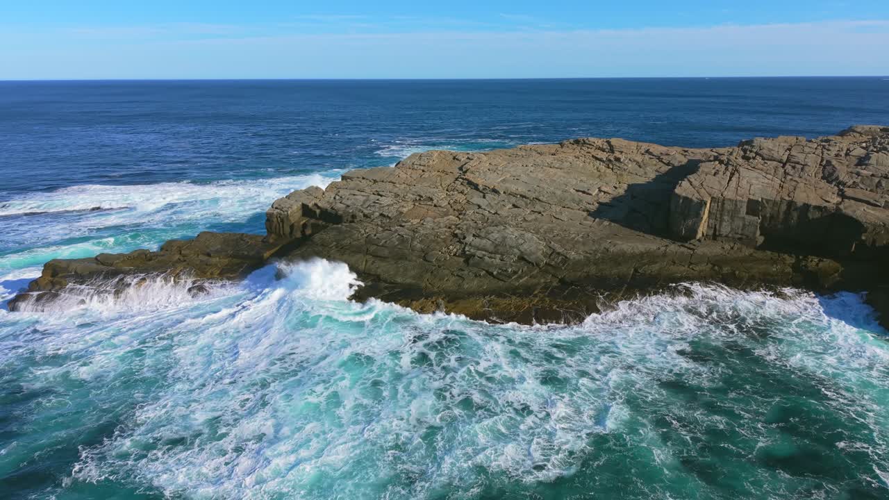 Dramatic aerial footage captures Flatrock Beamer in Newfoundland where powerful waves crash against rugged coastal rocks and spray bursts into the air under a clear blue sky