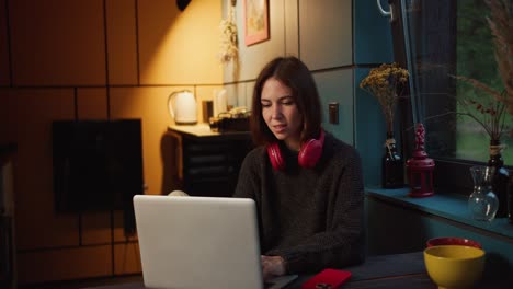 una feliz chica morena con auriculares rojos se regocija y mira la pantalla de una computadora portátil blanca en una habitación oscura de la noche iluminada por una lámpara amarilla