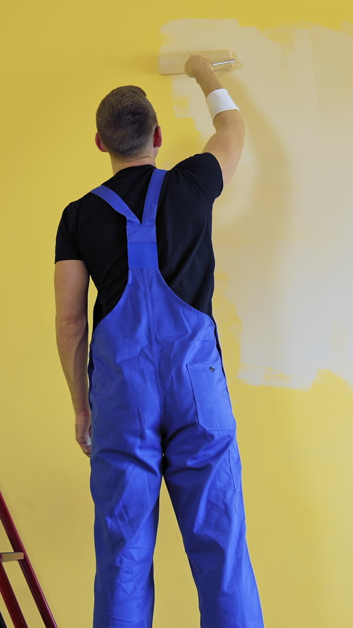 Painter doing the makeover indoors. Male worker painting wall with a paint roller next to the ladder. Improvement of a room interior. Back view. Vertical video