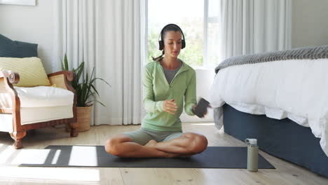woman in headphones sitting cross-legged on yoga mat, using smartphone at home