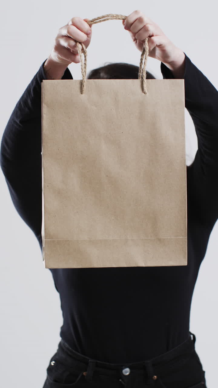 Vertical video of caucasian woman holding paper shopping bag with copy space on white background