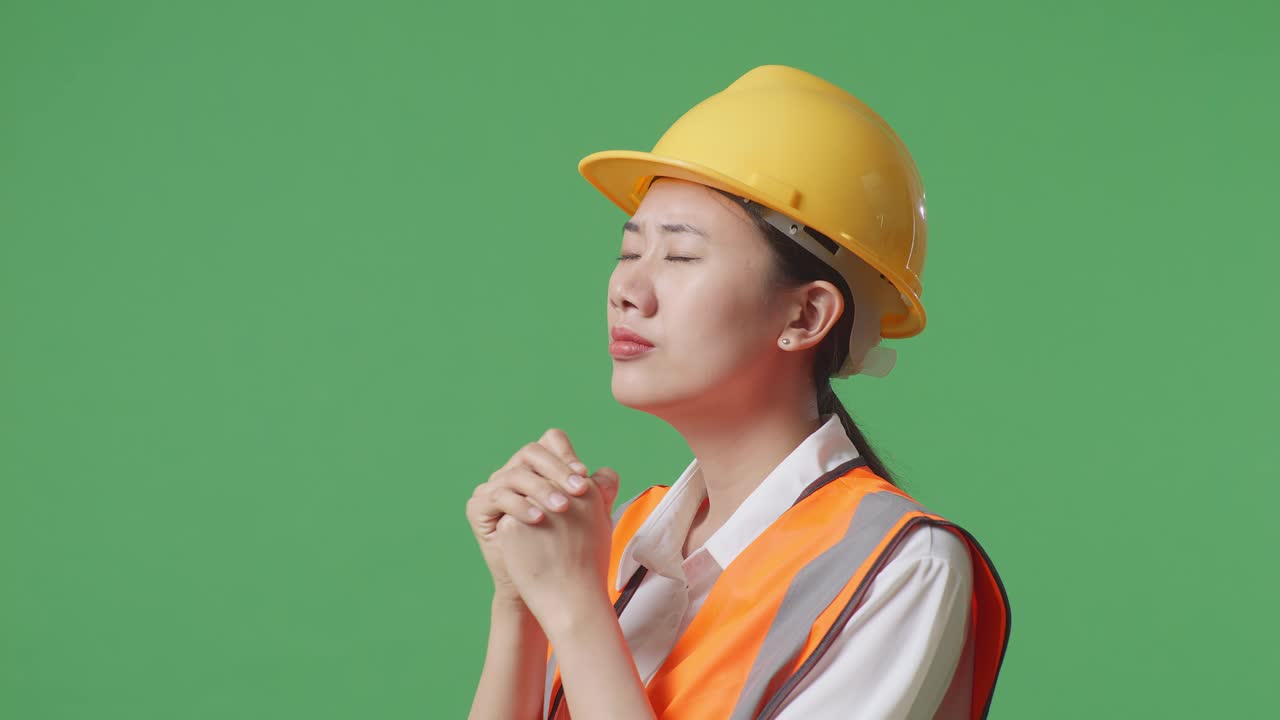 안전 헬을 입은 아시아 여성 엔지니어가 녹색 스크린 배경 스튜디오에 서있는 동안 무언가를 위해 기도합니다.