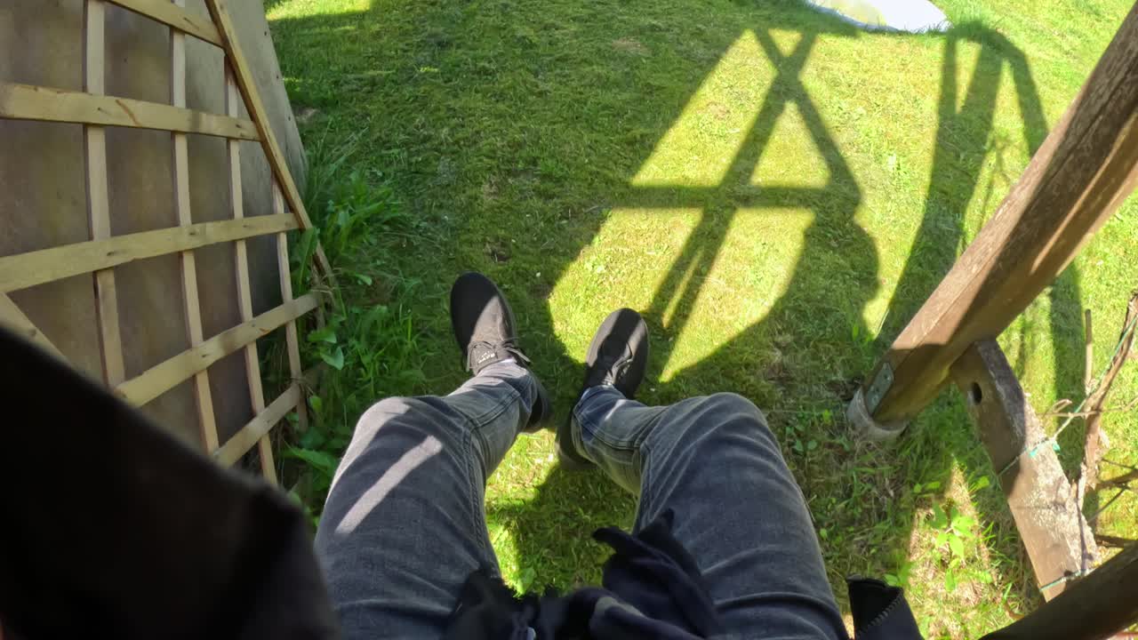 First-person perspective looking down at legs in dark jeans and black sneakers, dangling in a swing, above sunlit green grass with Shadows from an unseen structure above suggest height