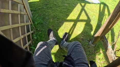 first-person perspective looking down at legs in dark jeans and black sneakers, dangling in a swing, above sunlit green grass with shadows from an unseen structure above suggest height