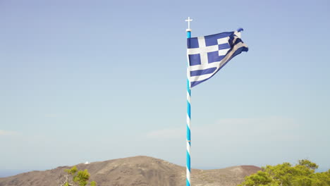 a greek flag blows in the wind with tall cliffs in the background