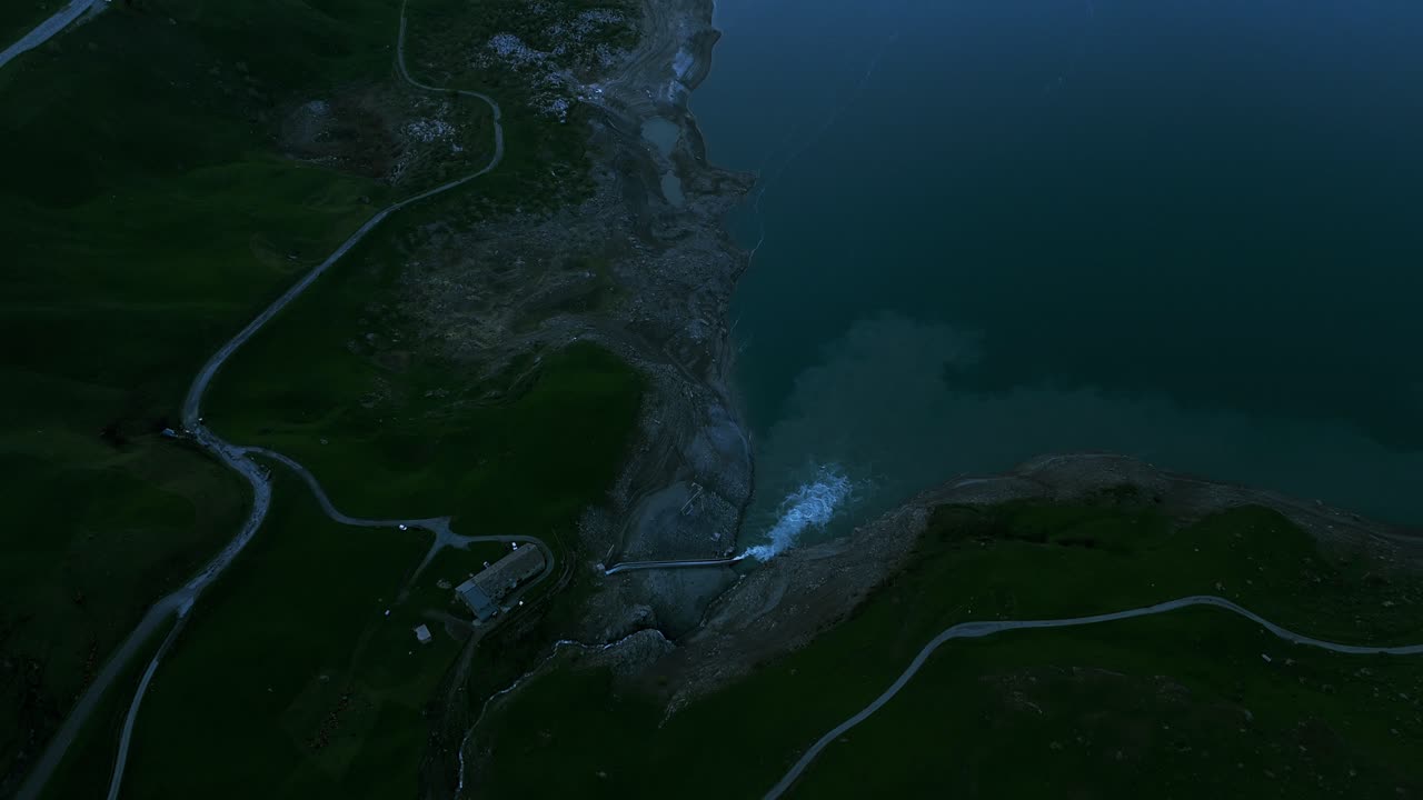 눈으로 인 높은 산들 사이에 자리잡은 평온한 호수의 공중 촬영, 해가 지는 동안 물 표면과 휘어지는 도로에서 반사되는 부드러운 빛으로 찍은