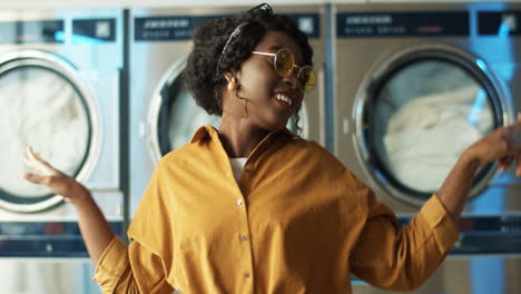 Pretty-Cheerful-Girl-In-Yellow-Glasses-Having-Fun-And-Dancing-In-Laundry-Service-Room-While-Machines-Washing-On-Background