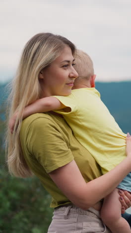 cheerful woman cuddles little boy holding son in arms against river running in ravine and dark mountains at highland on cloudy day slow motion