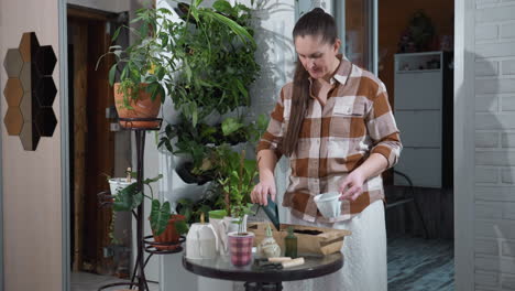 woman wearing plaid shirt stands by indoor table pouring soil into white planter cup with green scoop, focused expression, surrounded by thriving potted plants and gardening tools in calm setting