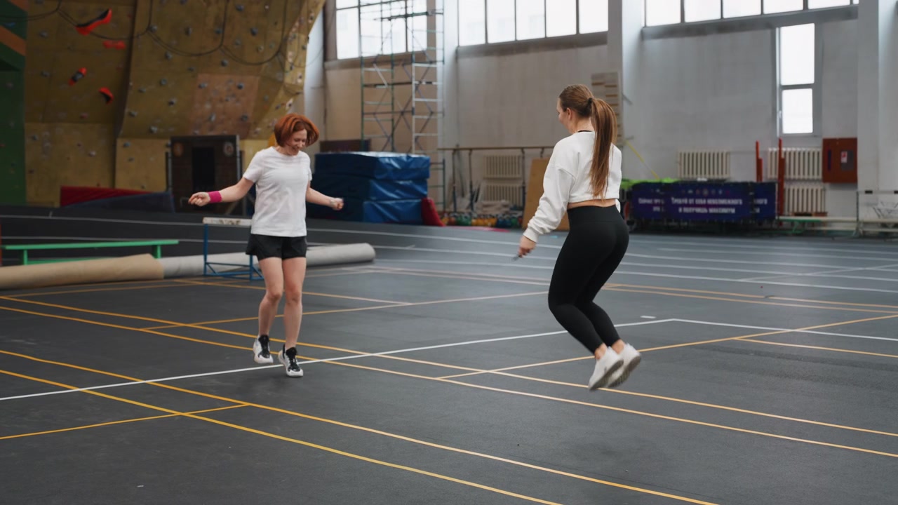 Due donne che praticano badminton in una palestra interna, scambio leggero e riscaldamento su un campo segnato, sessione di allenamento informale con volano e racchette, sorrisi e gioco di gambe cooperativo vicino a una parete da arrampicata e panchine.