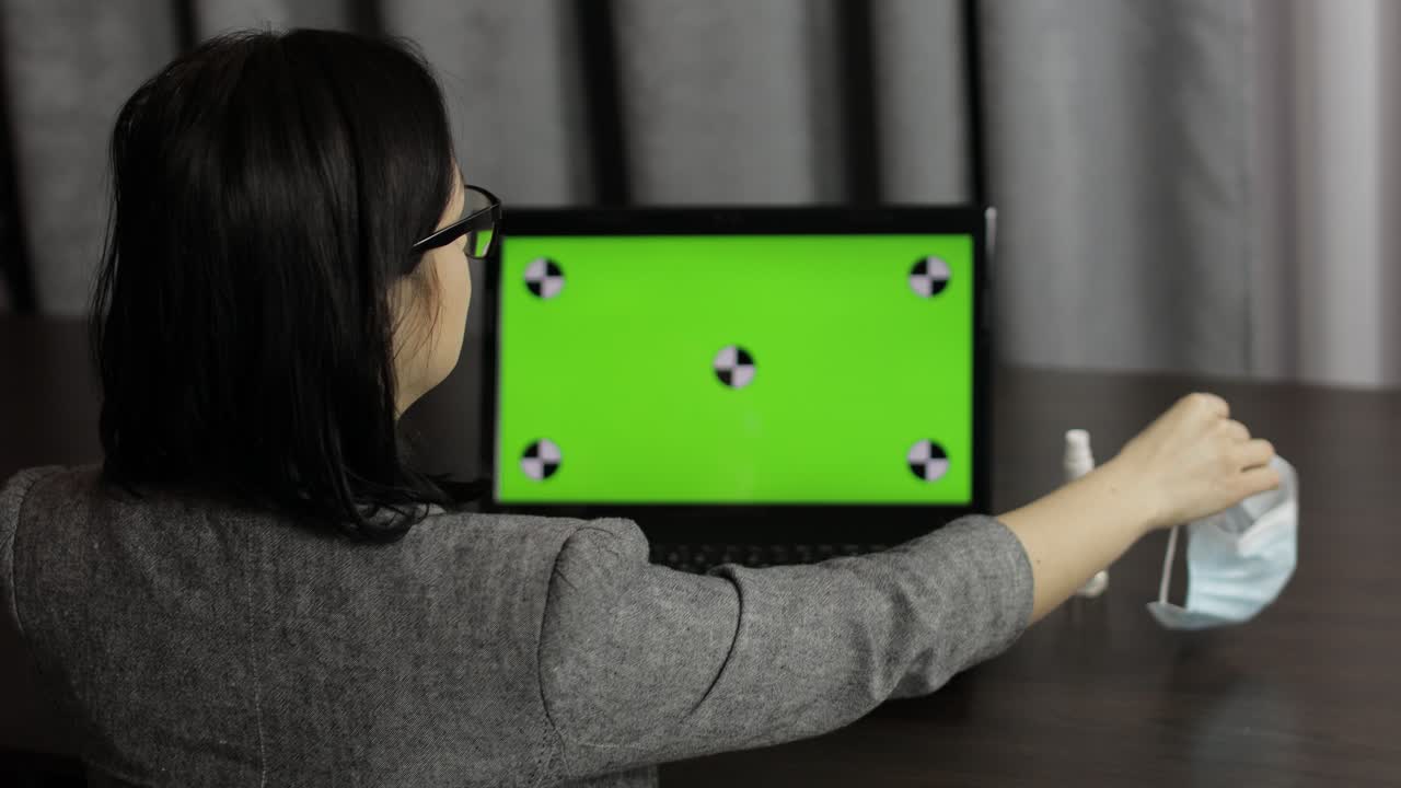 여성은 의료 마스크를 제거하고, 세니타이저를 가져와 녹색 화면의 노트북 근처에서 사용합니다.