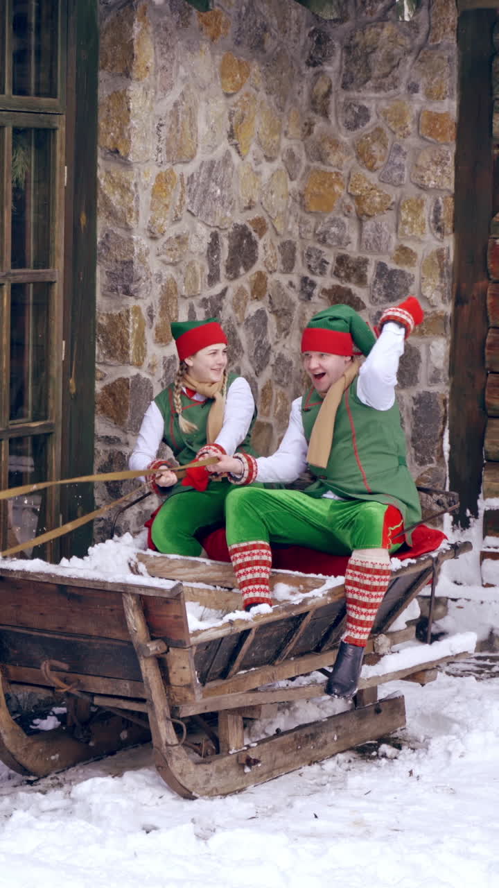 Cheerful elves having time together at Christmas outside in winter. Fairy characters sitting in Santa's sleigh on wooden building background. Vertical video