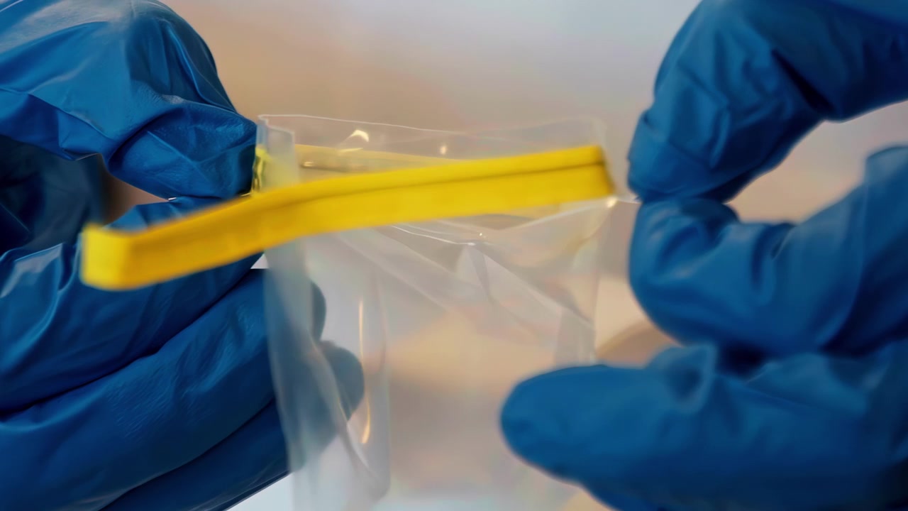 Mãos com luvas azuis abrindo uma bolsa plástica transparente selada com uma tira amarela.