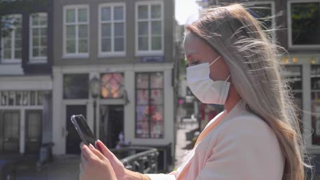 tourist woman taking selfie using her cellphone standing on canal bridge in amsterdam, netherlands during covid-19 outbreak