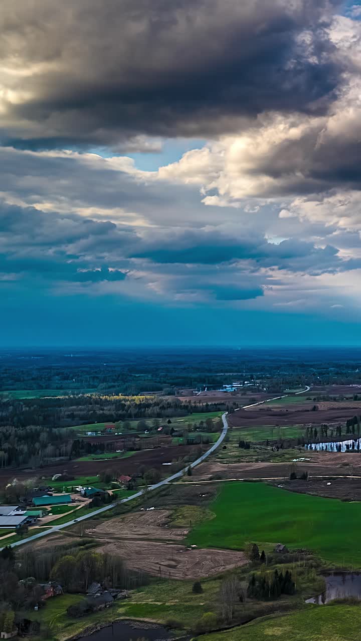 Timelapse aereo con drone sulla campagna lettone con una strada tortuosa che attraversa campi coltivati a mosaico, sotto drammatiche nuvole temporalesche e un cielo blu vivido.