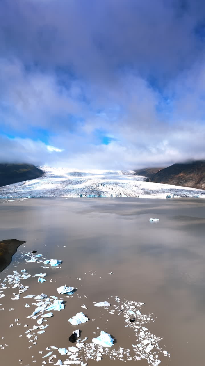 Iceland scenery from drone revealing view on beautiful glacier descending to the lake. Low angle view at grey clouds hiding the blue sky. Vertical video.